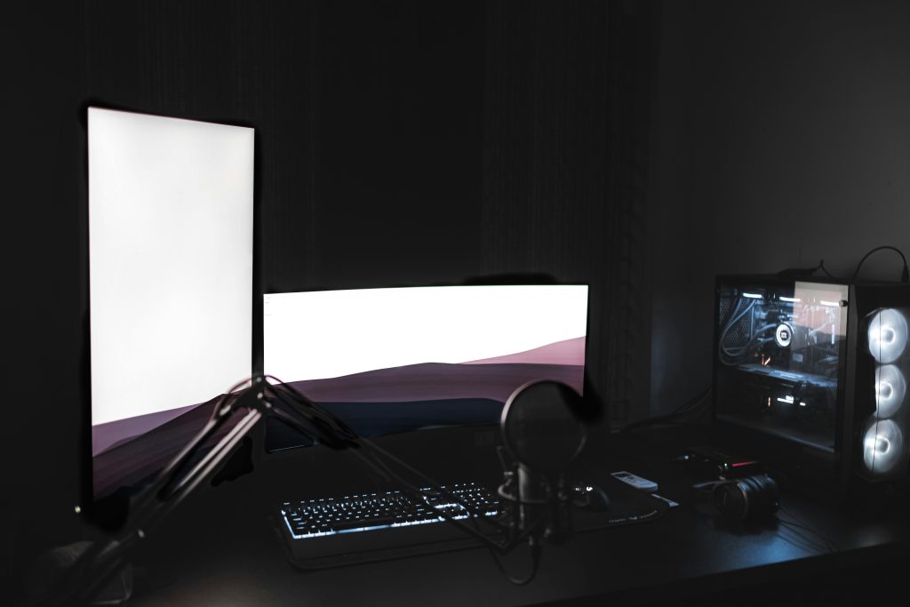 Gaming setup with glowing modern monitors placed on black table with keyboard near microphone and system unit in dark room